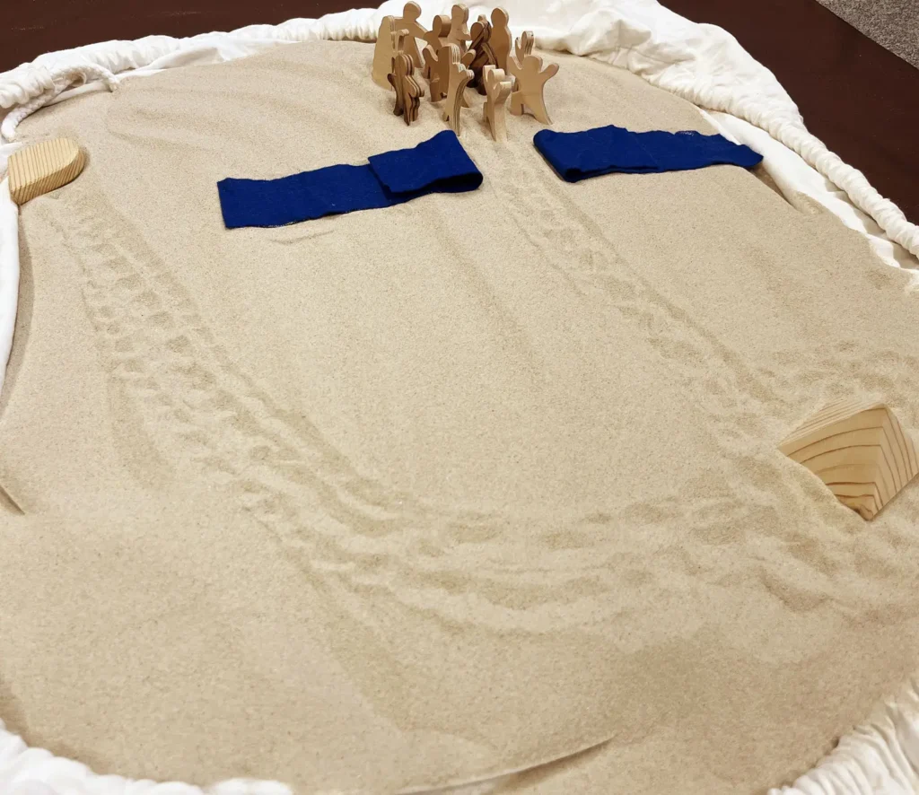 Foto eines Bodenbildes: Sand mit Spuren, Pyramide aus Holz, zwei blaue Tücher, die einen Streifen Sand freilassen, dahinter steht eine Gruppe von Holzfiguren.