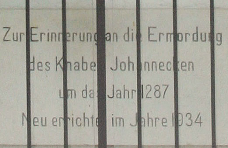 Ein Foto, durch Vergitterung fotografiert, von Eisenstäben durchbrochen, von dunkler Inschrift auf hellem Grund. Inschrift Johänneckenkapelle: Zur Erinnerung an die Ermordung des Knaben Johännecken um das Jahr 1287.