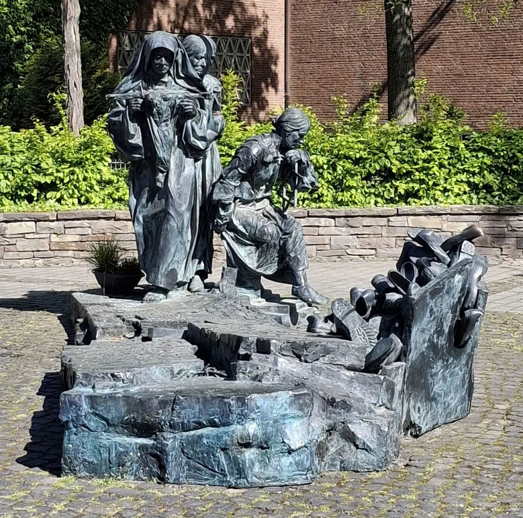 Ein Bronzedenkmal mit drei Frauenfiguren, links eine Ordensfau, dahinter eine stehende Frau, rechts sitzend eine Frau mit Davidstern. Im Vordergrund rechts ein Haufen von Schuhen.